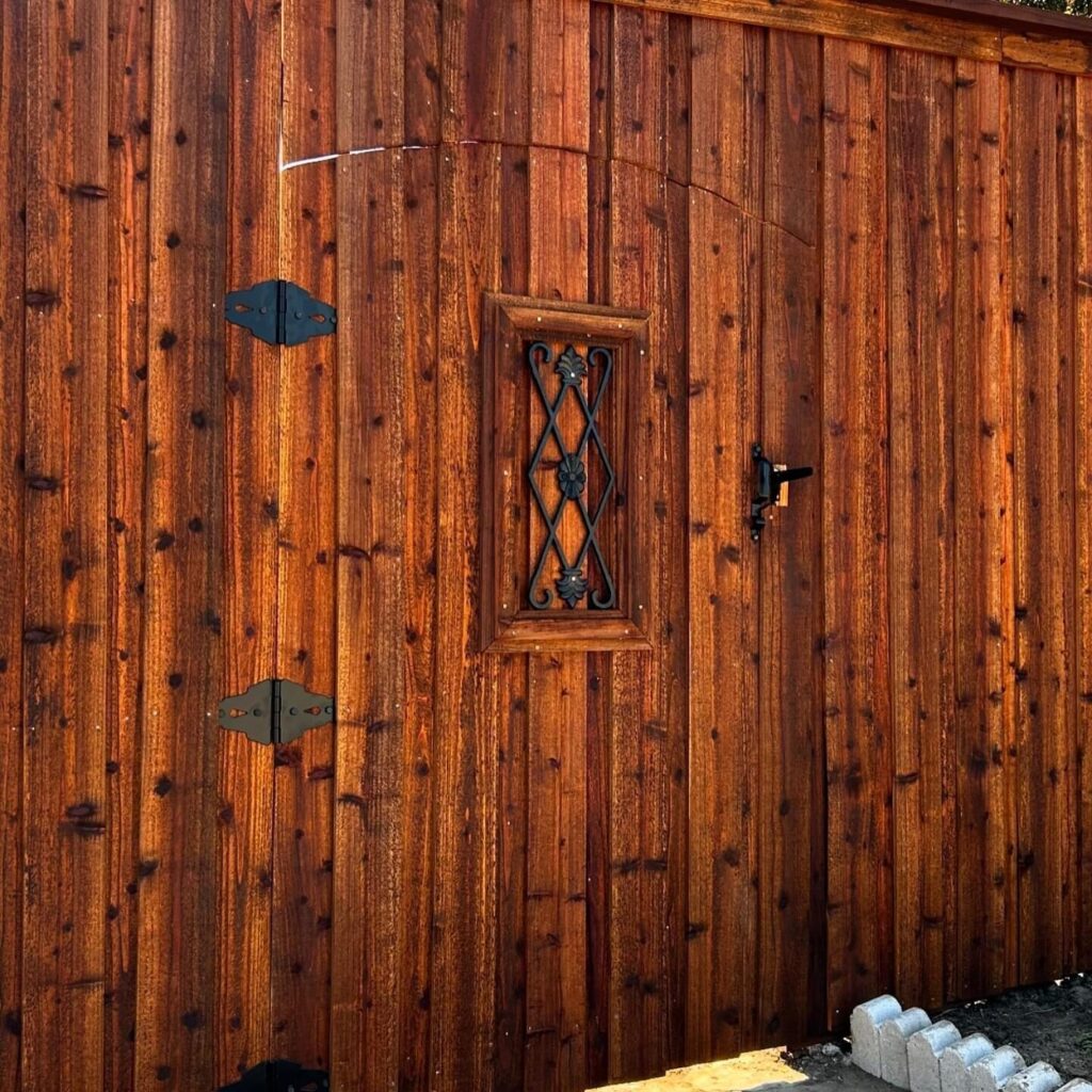 Close-up of a cedar gate with a wrought iron window and black strap hinges