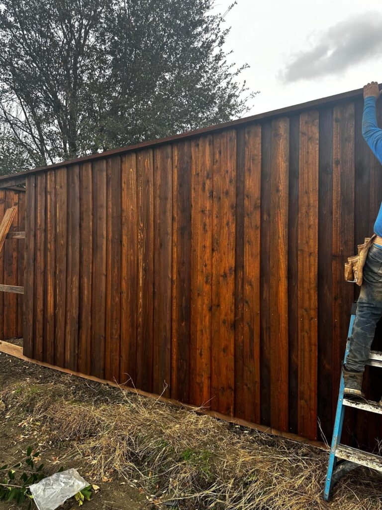 Grace Construction crew member finishing a stained cedar privacy fence from a ladder